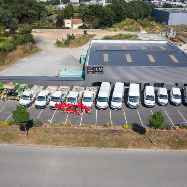 Equipe JCM Création Photographie aérienne des locaux de JCM Création. Le bâtiment professionnel gris est entouré d’une cour et d’un parking. Une importante flotte de véhicules utilitaires blancs et plusieurs engins de chantier sont stationnés devant les locaux. L’équipe de l’entreprise, vêtue majoritairement de rouge, est regroupée au centre du parking. Les enseignes JCM Création et Maisons du Choletais sont visibles sur la façade.